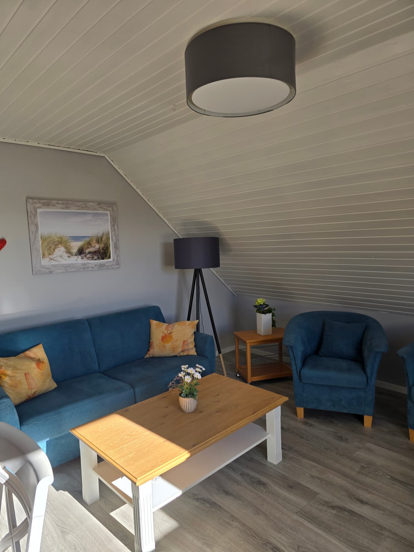 Wohnbereich mit blauer Couch, Sesseln, Stehlampe und Strandbild unter der Dachschräge